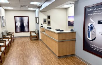 A brightly lit medical office reception area with a long wooden front desk. To the left, there is a row of gray upholstered chairs for patients. Large framed posters on the walls advertise robotic joint replacement surgery by Dr. Frederick F. Buechel, Jr., and several awards are displayed behind the counter.