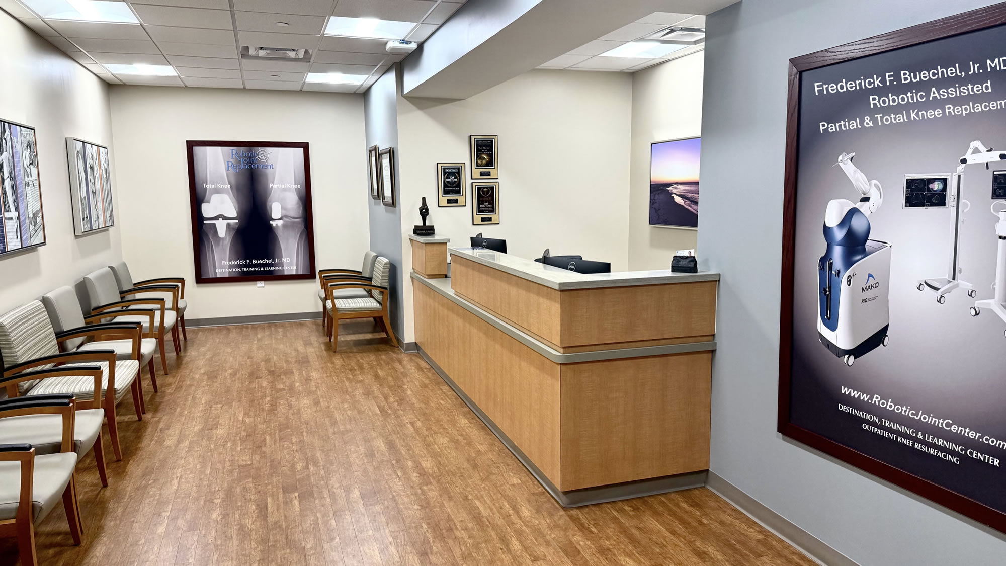 A brightly lit medical office reception area with a long wooden front desk. To the left, there is a row of gray upholstered chairs for patients. Large framed posters on the walls advertise robotic joint replacement surgery by Dr. Frederick F. Buechel, Jr., and several awards are displayed behind the counter.