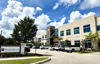 The exterior of a modern two-story medical building under a bright blue sky with fluffy white clouds. A sign in the foreground reads "Physicians Regional" next to the address 8831. The parking lot in front of the beige and glass building is filled with several cars and SUVs.