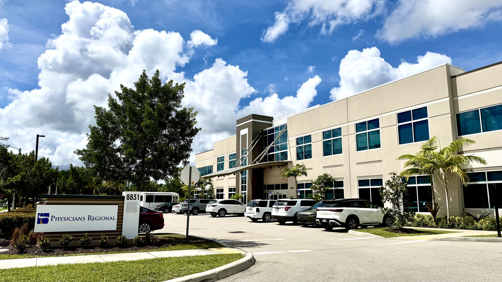 The exterior of a modern two-story medical building under a bright blue sky with fluffy white clouds. A sign in the foreground reads "Physicians Regional" next to the address 8831. The parking lot in front of the beige and glass building is filled with several cars and SUVs.