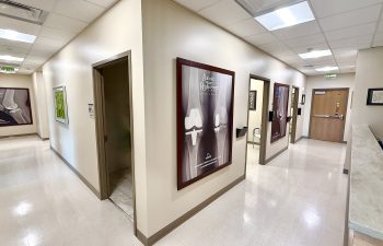 A bright, sterile hospital hallway with white tile floors and cream-colored walls. Large framed educational posters about knee replacements are mounted along the corridor. The hallway branches off to various examination rooms and a secondary reception desk is visible on the right.