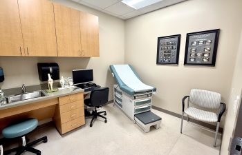 A clean, modern medical examination room featuring a blue and white exam table with a paper cover. The room includes a wooden workstation with a computer, a stainless steel sink, and anatomical models of a knee joint. On the wall, two framed posters display detailed knee anatomy and X-ray images.