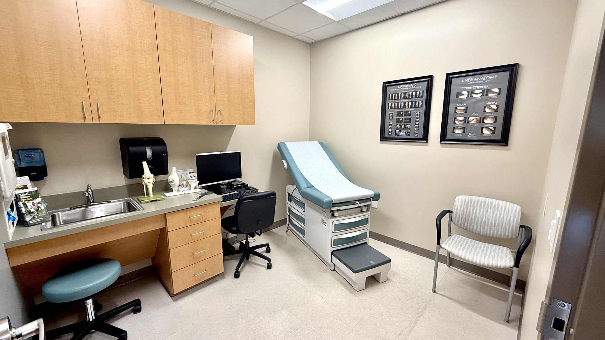 A clean, modern medical examination room featuring a blue and white exam table with a paper cover. The room includes a wooden workstation with a computer, a stainless steel sink, and anatomical models of a knee joint. On the wall, two framed posters display detailed knee anatomy and X-ray images.