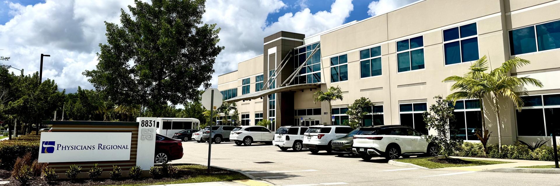 The exterior of a modern two-story medical building under a bright blue sky with fluffy white clouds. A sign in the foreground reads "Physicians Regional" next to the address 8831. The parking lot in front of the beige and glass building is filled with several cars and SUVs.