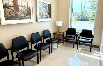A small, sunlit waiting nook with five black upholstered chairs arranged against the walls. A large window overlooks a parking lot, and two framed photographs of nature and a bicycle hang on the wall. A small wooden side table with a lamp sits in the corner.
